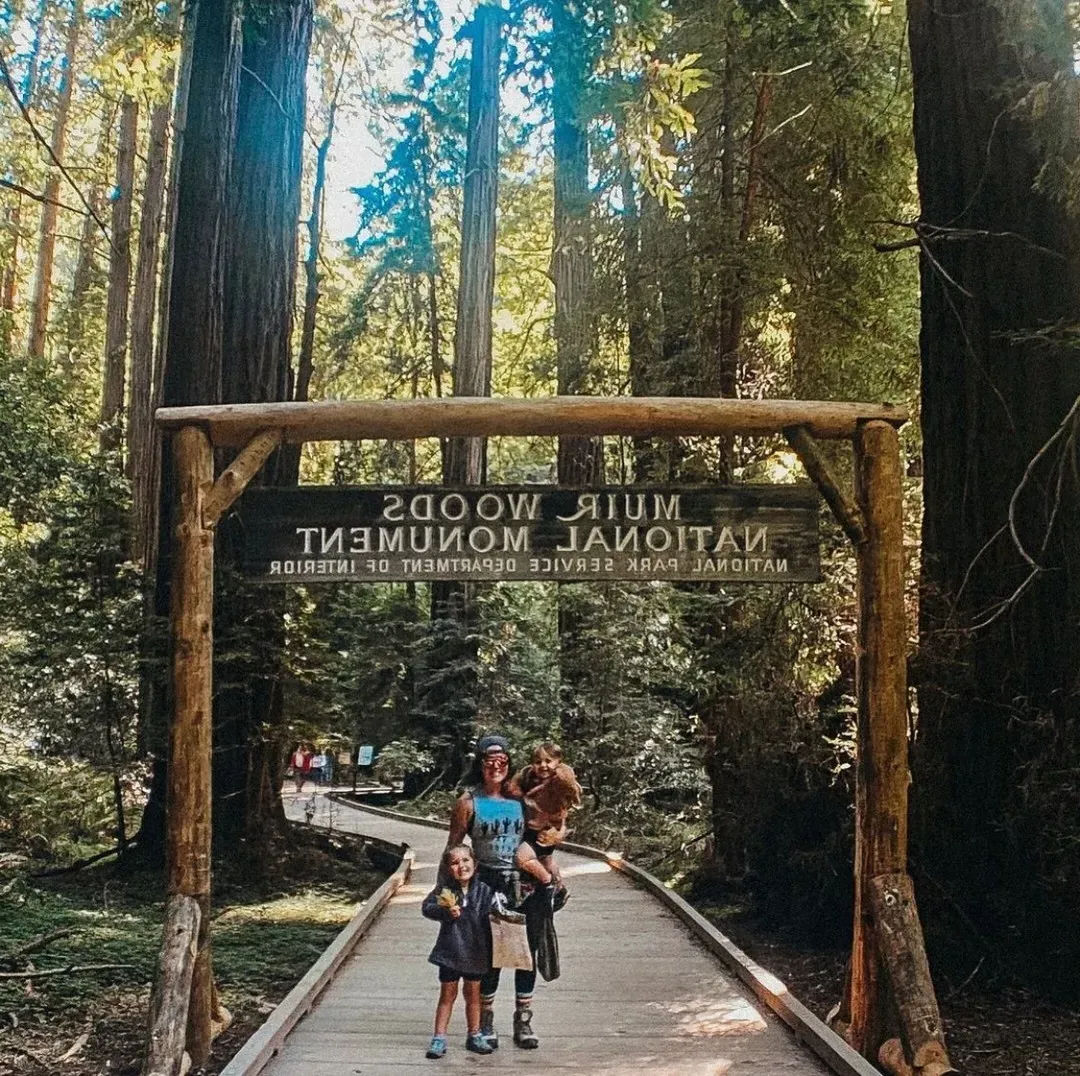 《妇女与儿童站在Muir Woods国家纪念碑标志前，在森林的一条木制小径上